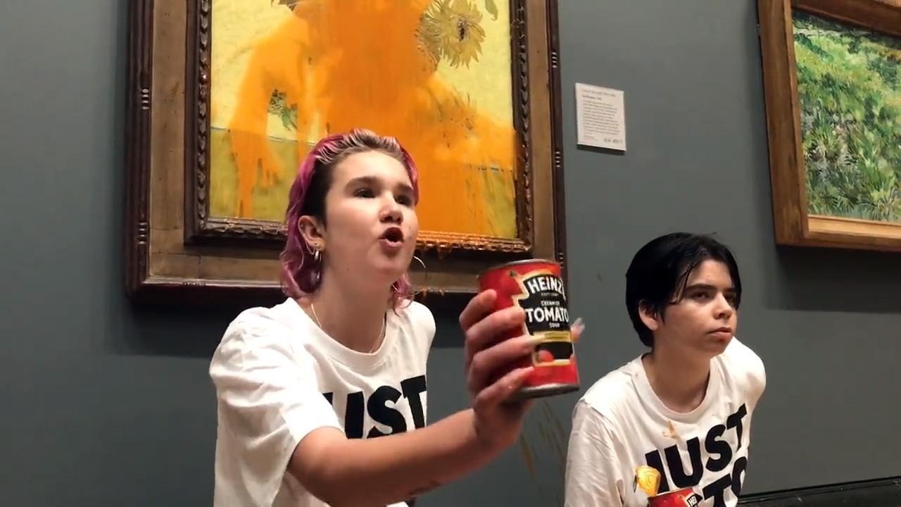 Two young people, one with pink hair, the other with dark brown hair, kneel in front of Sunflowers, its glass covered with dripping red soup. The pink haired protester holds up a Heinz Tomato soup can and appears to be speaking.